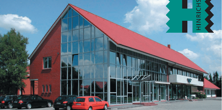 Modernes, mehrstöckiges Gebäude mit einer großen Glasfassade, rotem Satteldach und weißen Fassadenteilen. Im Vordergrund sind mehrere parkende Fahrzeuge, darunter rote und schwarze Autos. Der Himmel ist teilweise bewölkt.