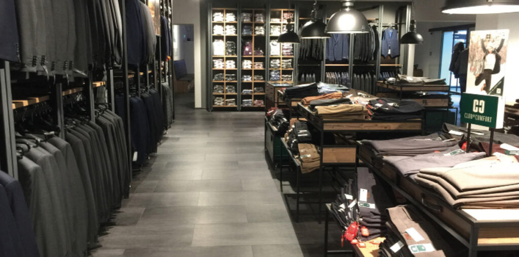 Blick in einen Bekleidungsgeschäftsraum mit dunkel gefliesten Boden, Regalen und Tischen, die Hemden, Hosen, Jacken und andere Kleidungsstücke anzeigen. Kleidung ist ordentlich angeordnet. Es gibt beleuchtete Aufsteller im Hintergrund.