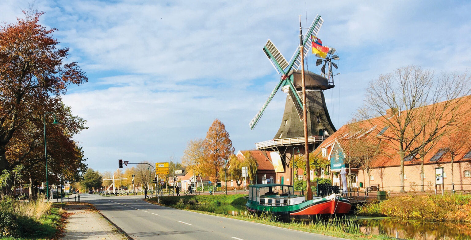 Das Bild zeigt eine holländische Windmühle neben einem Kanal, umgeben von Bäumen mit Herbstfärbung und einem Straßenzug mit Verkehrsampel. Die Windmühle hat vier Flügel, trägt eine niederländische Flagge und steht vor einem roten Backsteinhaus. Der Himmel ist teilweise bewölkt.