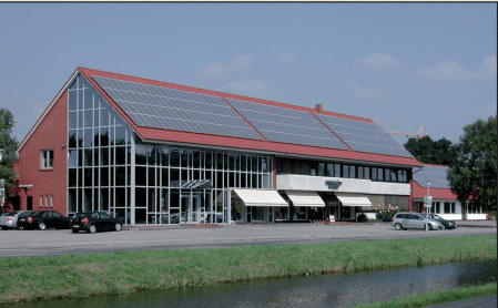 Modernes, mehrstöckiges Gebäude mit einer großen Glasfassade, rotem Satteldach mit Solarpanelen, mehreren Eingängen mit weißen Sonnenschutzmarkisen und umliegenden Parkplätzen; dahinter Bäume und blauer Himmel.