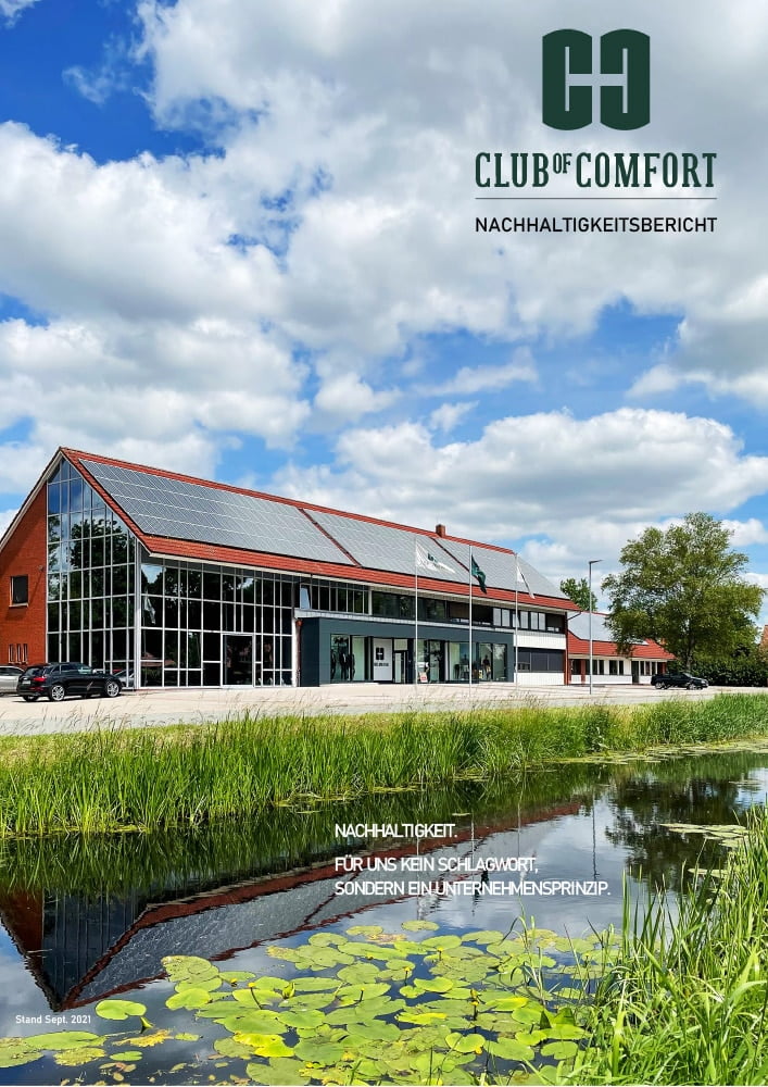 Moderne, zweistöckige Unternehmenszentrale des CLUB OF COMFORT mit großen Glasfassaden, Solarpanelen auf dem Dach und einem Parkplatz im Vordergrund. Im unteren Bereich befindet sich ein kleiner Teich mit Seerosen und grüner Vegetation. Über dem Gebäude steht das grüne Logo des CLUB OF COMFORT und der Schriftzug "NACHHALTIGKEITSBERICHT". Das Bild wurde im September 2021 aufgenommen.