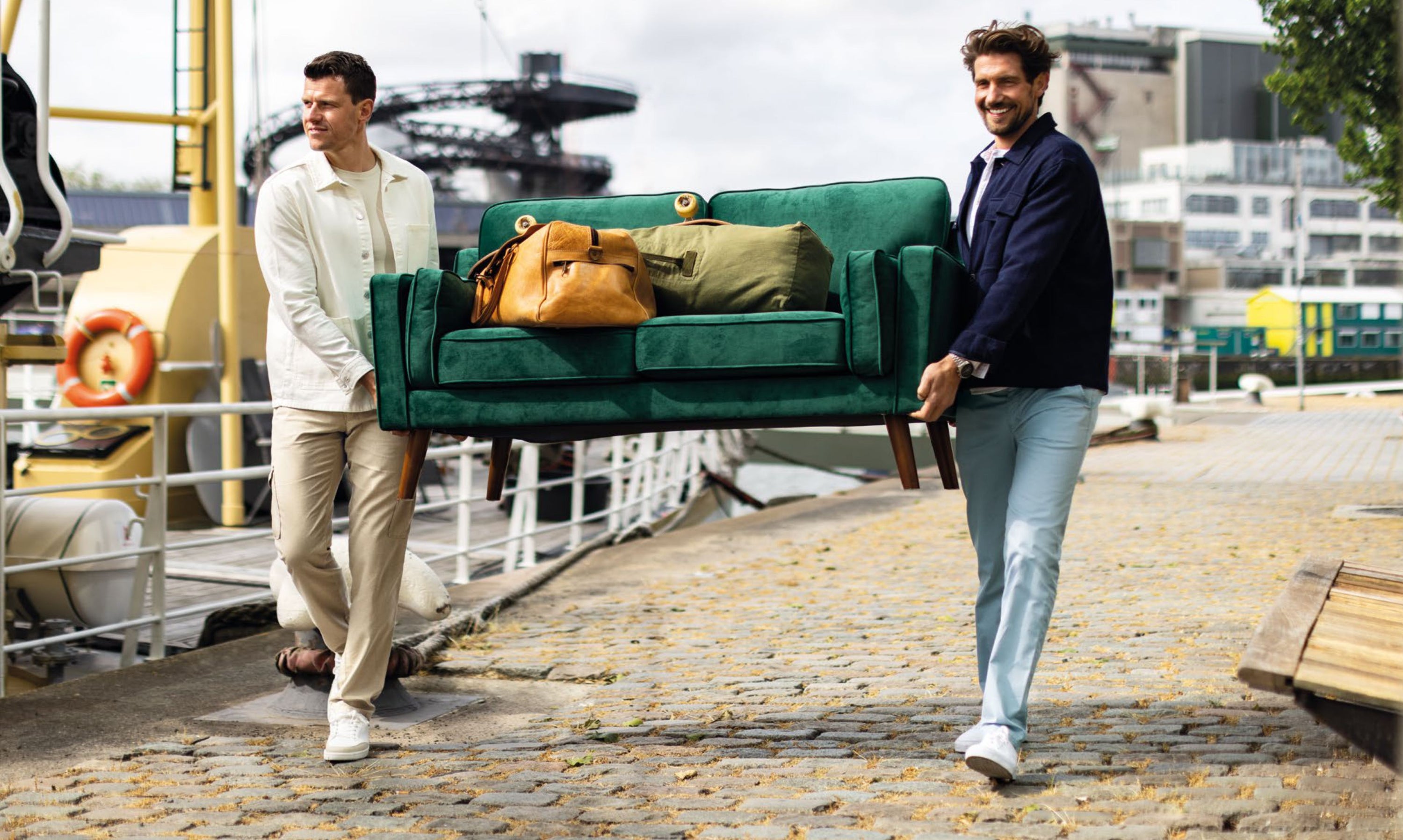 Zwei Männer tragen einen grünen Sofa mit einigen Taschen darauf, während sie entlang eines Hafenbeckens auf einer gepflasterten Promenade laufen. Im Hintergrund sind Schiffe, Gebäude und ein Kran sichtbar.