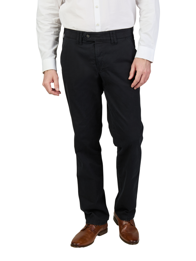 Porträt eines Mannes, der schwarze Anzugshosen, braune Lederschuhe und eine weiße Hemd trägt, sichtbar bis zur Taille. Seine Hände ruhen locker an den Seiten.