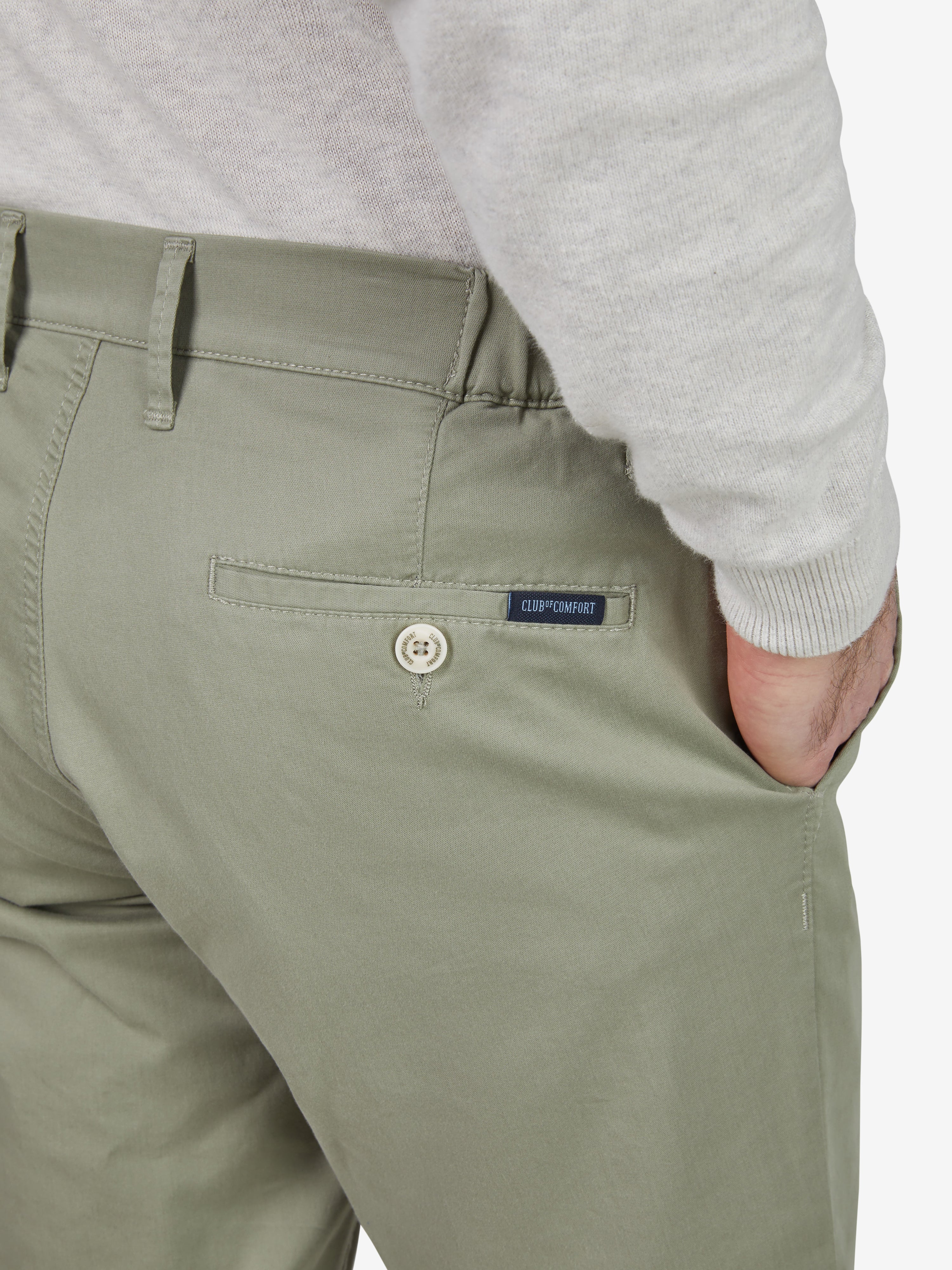 Nahaufnahme einer khakifarbenen Chinohose von Club of Comfort mit schmalem Bein, Comfort-Flex-Bund, Hemdenstopper, aus sommerleichter High Stretch Baumwolle mit 280 g/m², sichtbare Rücken-Reißverschlusstasche mit Label, getragen von einer Person in grauem Oberteil.