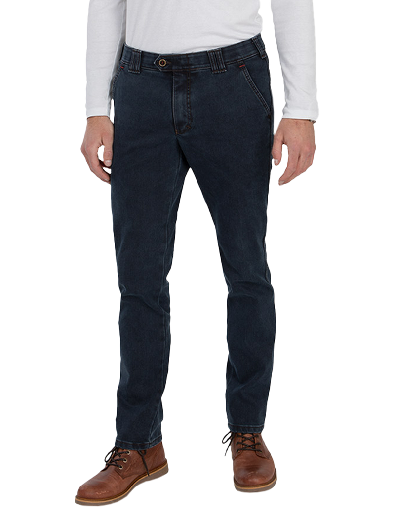 Ein Mann trägt dunkle Jeans, ein weißes langarmiges Oberteil, braune Lederstiefel und steht gegen einen weißen Hintergrund.