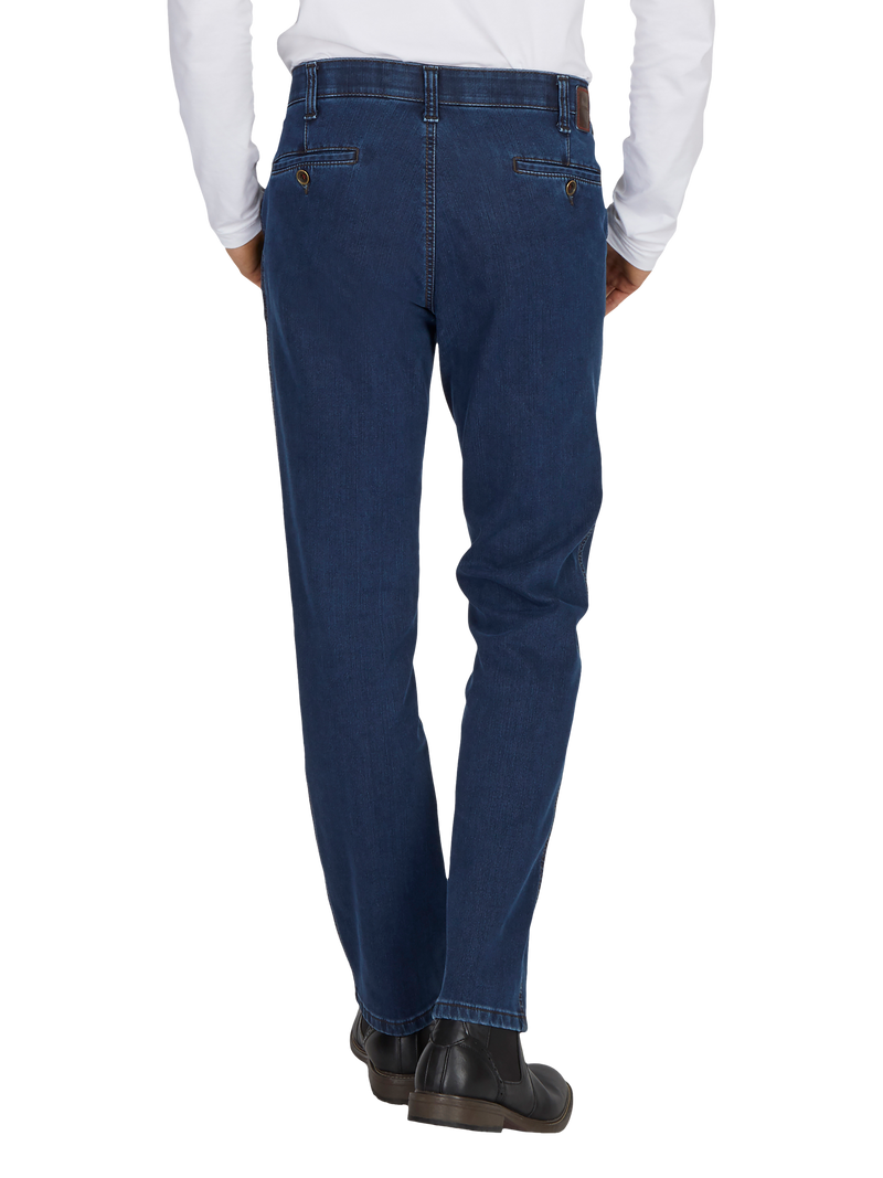 Ein Mann trägt eine dunkelblaue Jeanshose, die an der Taille sitzt, mit sichtbaren Nieten an den Ecken der Tasche und einem böhmischen Schnitt. Er trägt schwarze Schuhe und ein weißes, langarmiges Hemd, das aus der Hose herausschaut.