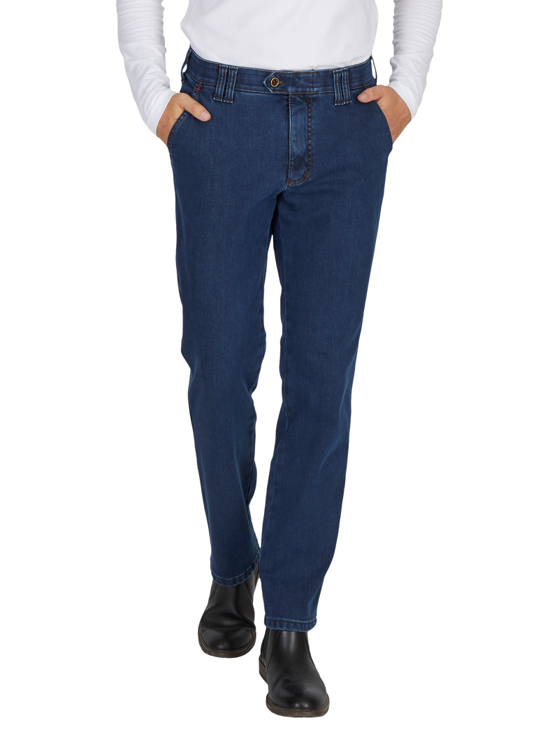 Nahaufnahme eines Menschen mit dunklen Jeans, die an der Taille sitzen und in den Taschen ruhen, sowie schwarzen Stiefeln. Die Person trägt eine weiße, langärmlige Bluse. Die Bilderfassung zeigt den unteren Brustbereich bis zu den Füßen.