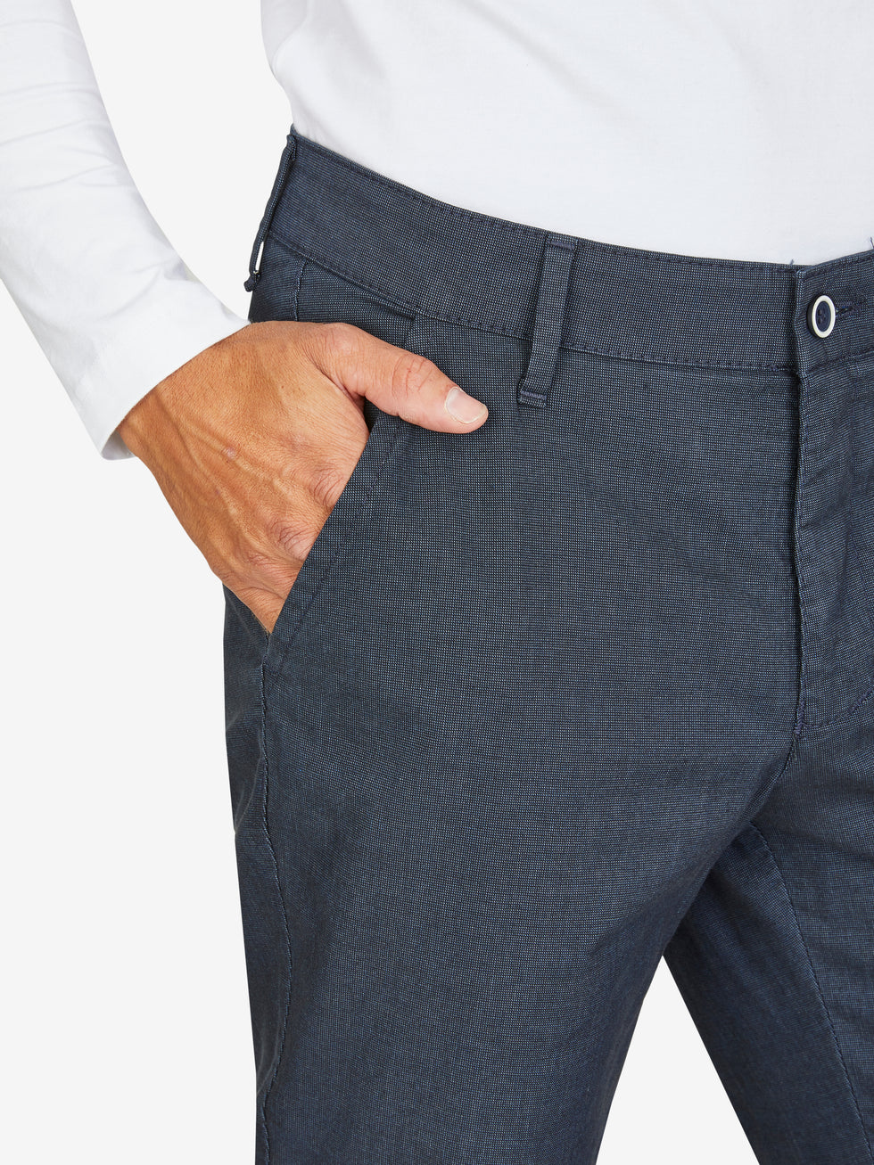 Nahaufnahme einer dunkelblauen, elastischen Flat Front-Hose mit Baumwoll-Mikrostruktur in Woll-Optik, mit Gürtelschlaufen und einer Hand im Bund. Modell zeigt Regular Fit, schmaler Beinverlauf, geeignet für Frühling/Sommer.