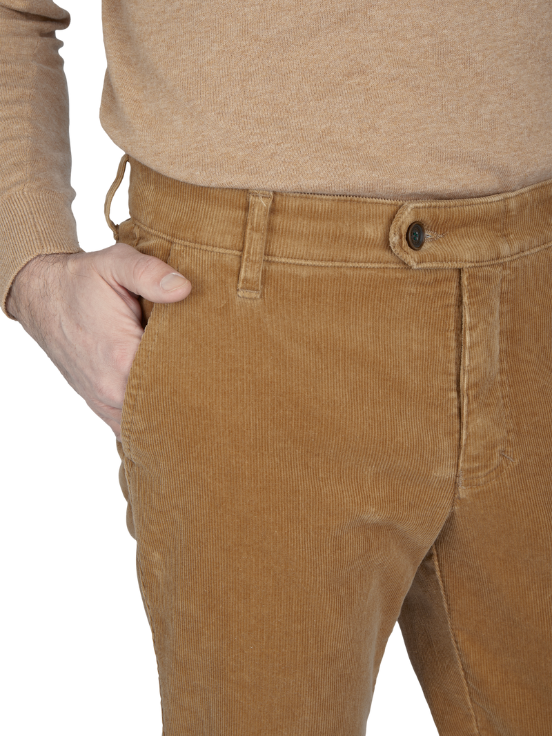 Ein Mann trägt braune Cordhosen und ein beige langärmeliges Oberteil. Seine rechte Hand ist in der Hosentasche.