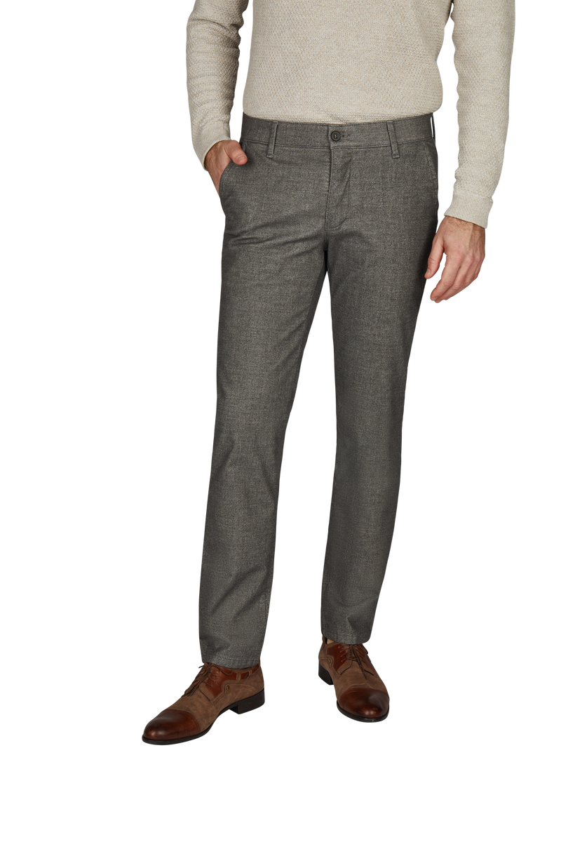 Herren trägt graue, formelle Anzugshosen, die vorne eine Reißverschluss- und Knopföffnung haben. Seine rechte Hand steckt in der Hosentasche, die linke hängt entspannt an der Seite. Er trägt braune Lederschuhe und einen hellen Pullover.