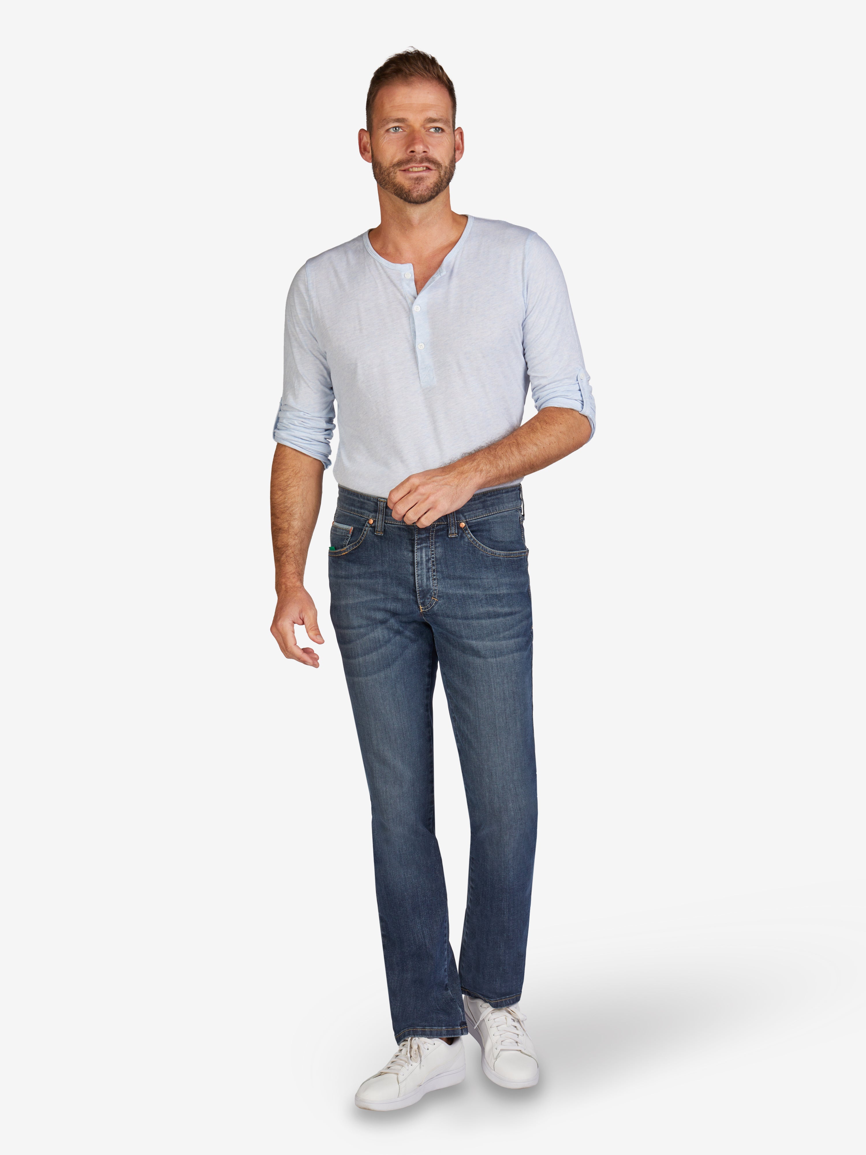 Mann trägt blaue, gewaschene HENRY 7915 6-Pocket Jeans mit elastischem Bund, Hemdstopper und Mobile Pocket, gefertigt aus hoch elastischem Super High Stretch-Denim, regulärer Fit, in einem hellen Waschlook, kombiniert mit weißem Hemd und Sneakers.