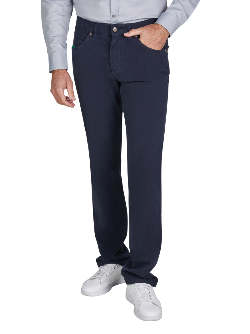 Ein Mann trägt dunkle Jeans, einen hellgrauen Hemd, weiße Sneakers und hat eine Hand in der Hosentasche.