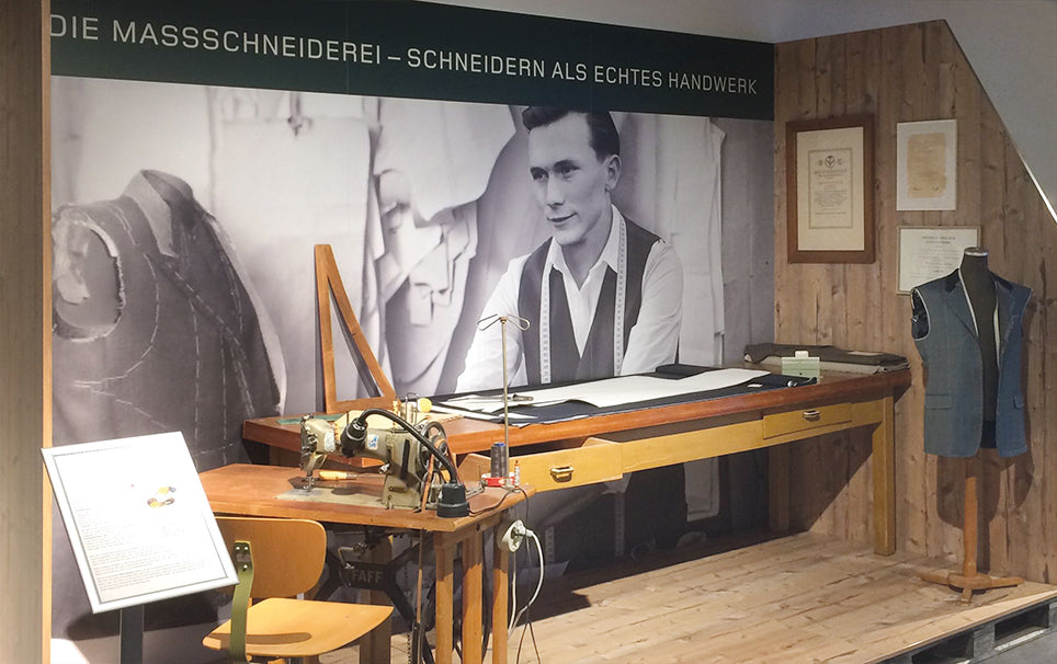 Historische Ausstellung mit einem Nähmaschinen-Arbeitsplatz, einem Sitzstuhl, einem Nähmaschinenmodell und einer Schaufensterpuppe mit Maßkleidung. Im Hintergrund ein Poster mit einem Mann und der Text „Die Massschneiderei – Schneidern als echtes Handwerk“.