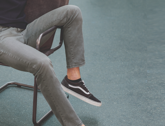 Ein Mensch sitzt auf einem Stuhl, die Beine sind im Überkreuzten auf einem Tisch, der sich außerhalb des Bildes befindet. Die Person trägt graue Hosen, schwarze Vans-Sneaker mit weißer Sohle, und der Hintergrund ist ein blauer, glatter Boden.
