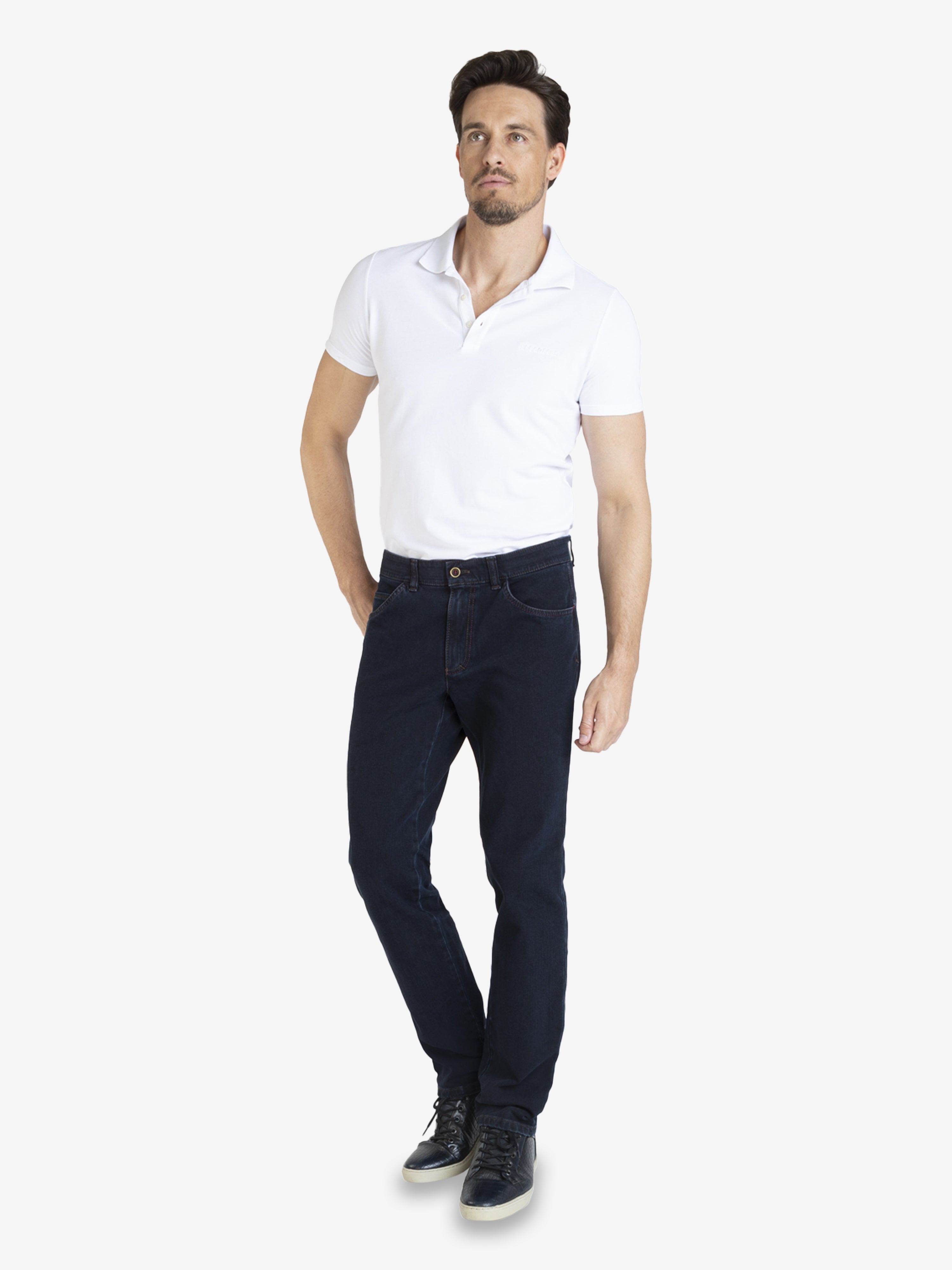 Mann trägt dunkelblaue MARVIN 7054 Jeanshose mit Regular Fit, elastischem Comfortbund, Swing-Pockets, aus hochelastischem Bi-Stretch-Denim mit T400-Beimischung, formstabil, inklusive Hemdstopper, model mit weißem Polo-Shirt, schwarze Schuhe.