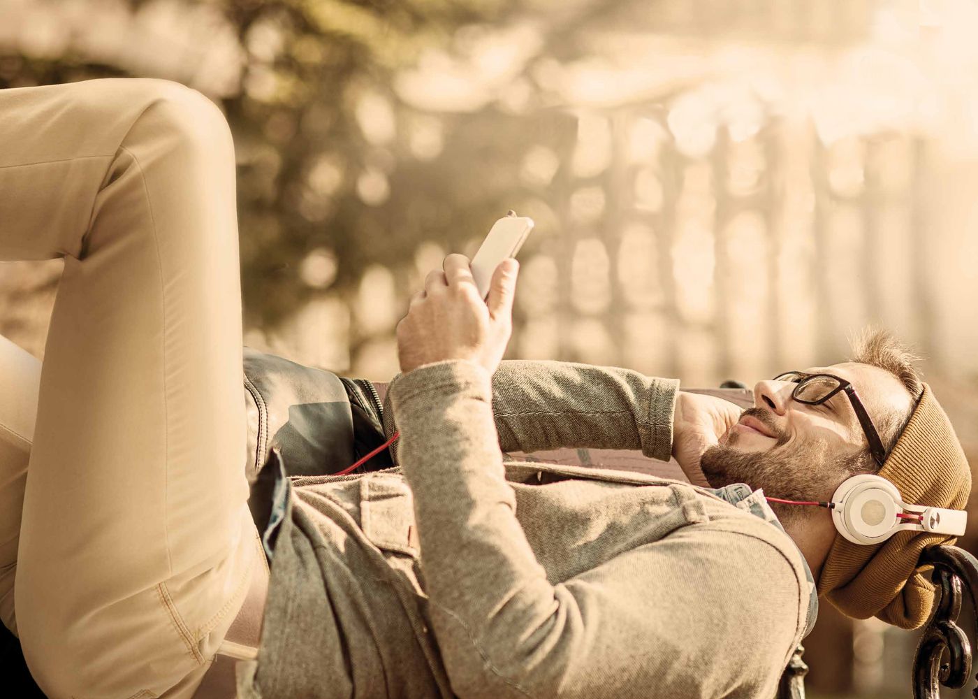 Ein junger Mann liegt entspannt im Freien auf einer Liege, trägt eine beige Mütze, Kopfhörer und Brille, und liest oder hört Musik auf seinem Smartphone. Im Hintergrund sind Bäume und Sonnenlicht zu erkennen.
