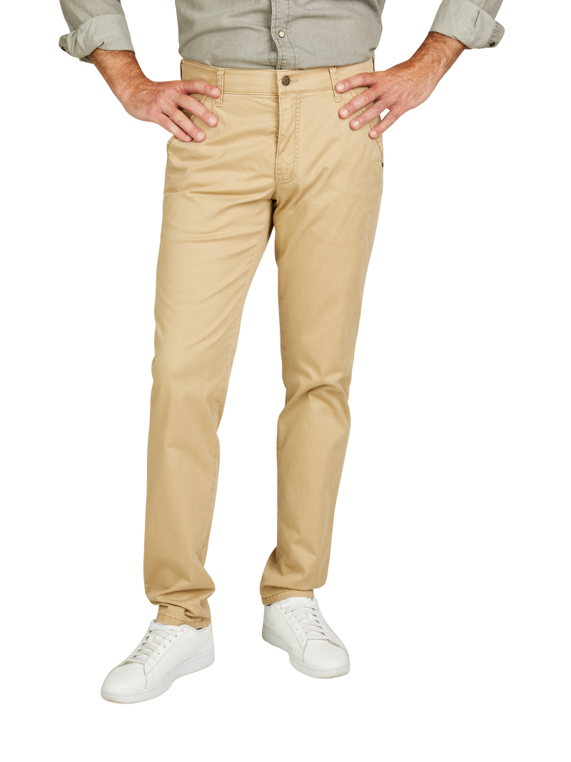 Der Bild zeigt einen Mann, der beige Jeans trägt, ein langärmeliges, hellgraues Hemd mit hochgekrempelten Ärmeln trägt, weiße Sneakers an den Füßen hat und seine Hände in die Taille steckt.