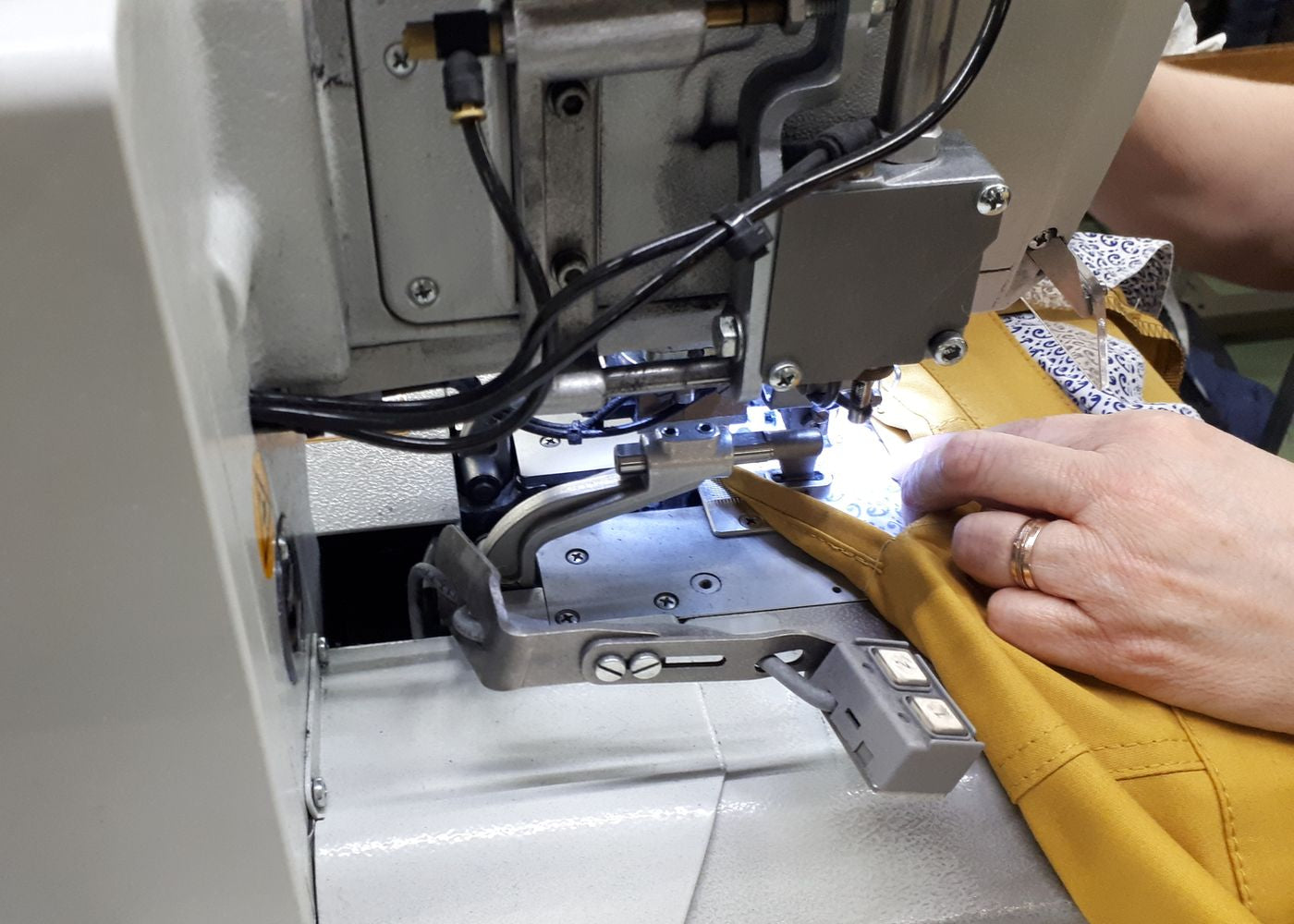 Nahaufnahme einer industriellen Nähmaschine, bei der eine Person mit beiden Händen einen goldgelben Stoff präzise führt, während die Maschine näht. Das Bild zeigt technische Komponenten, Kabel und die Fokusbildung auf den Nähvorgang.