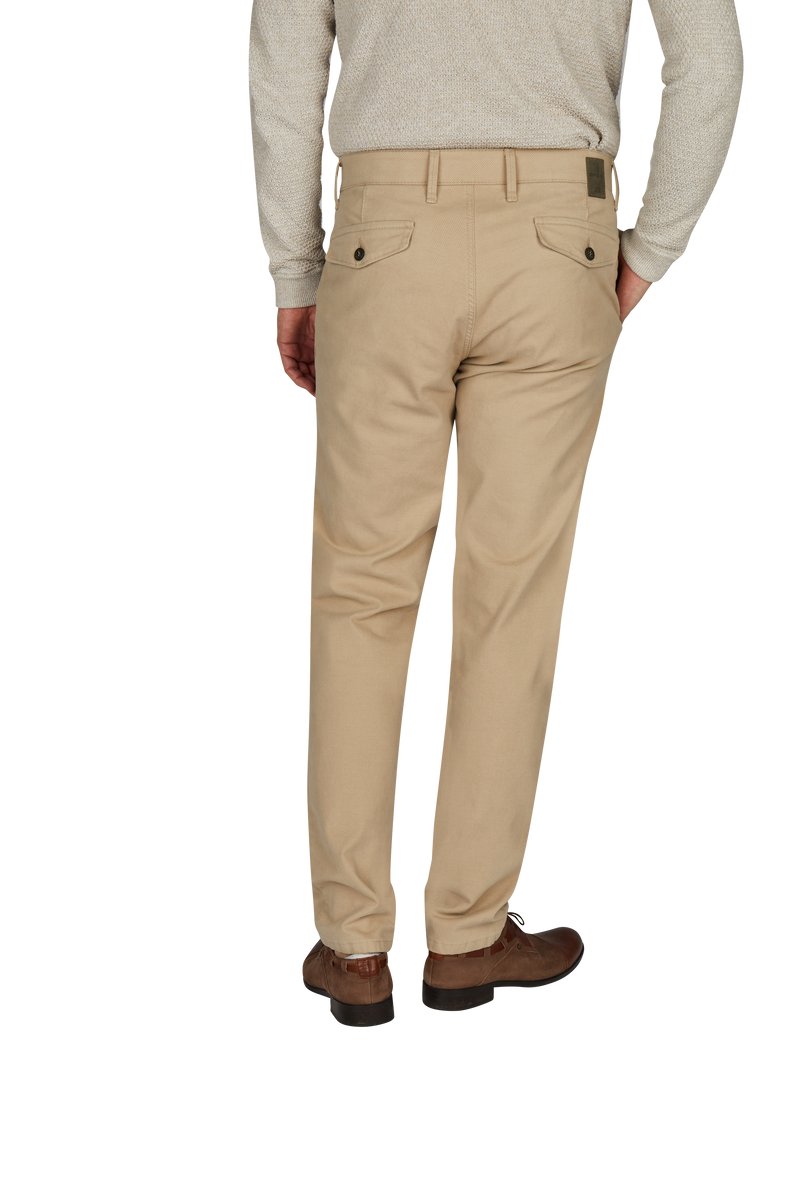 Ein Mann trägt beige Chinos, ein hellgraues Hemd oder Pullover, braune Schuhe und steht mit seiner rechten Hand in seiner Hosentasche, während er nach hinten blickt.