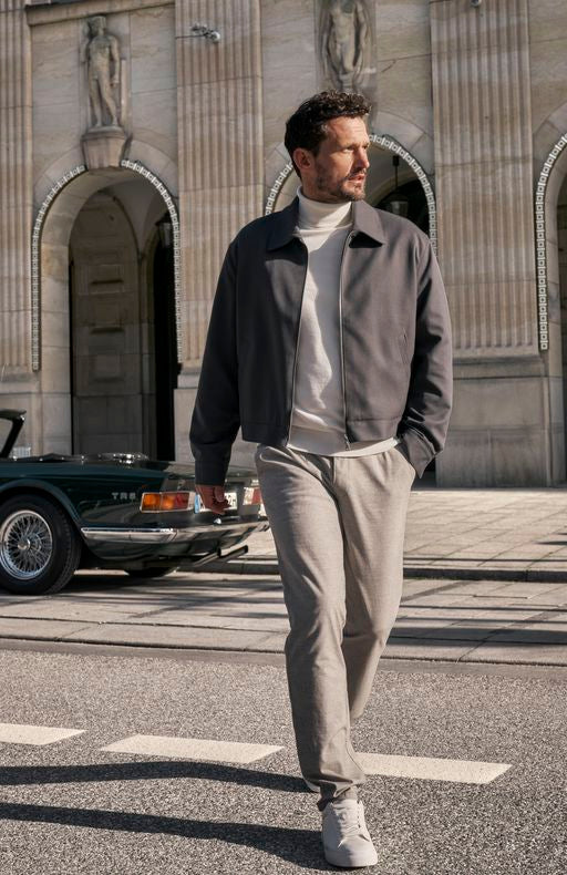 Ein Mann mit dunklen, kurzen Haaren und Bart läuft entspannt auf der Straße vor einer historischen Gebäude mit Statuen und Bögen. Er trägt einen grauen Jacke, ein helles T-Shirt, graue Hosen und weiße Sneakers. Im Hintergrund ist ein schwarzer Oldtimer zu sehen. Es ist ein sonniger Tag.