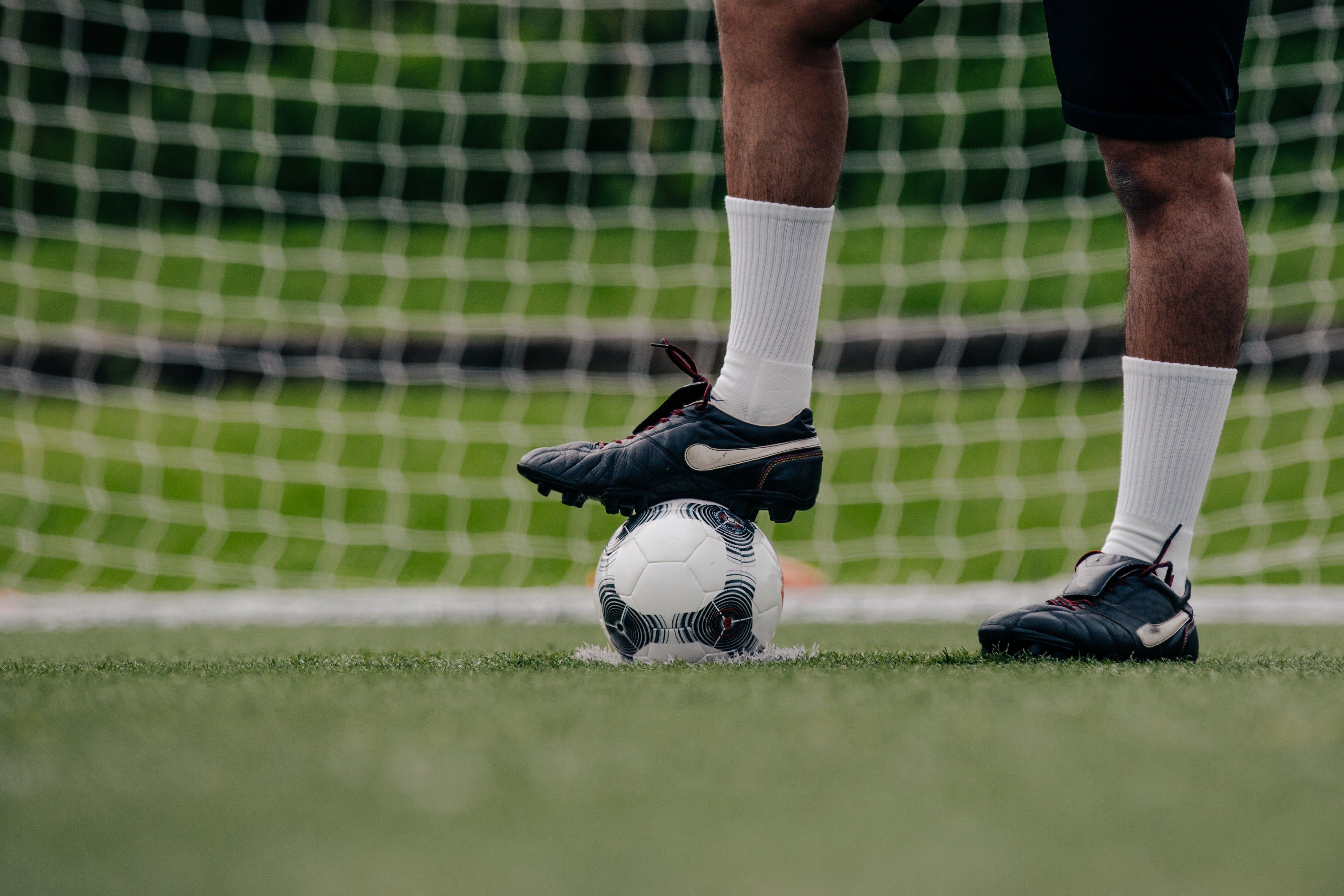 Nahaufnahme der Füße eines Fußballspielers, der auf einem grünen Rasen mit schwarzen Fußballschuhen und weißen Socken einen Fußball vor einem Tor kontrolliert. Im Hintergrund ist das Tornetz sichtbar.