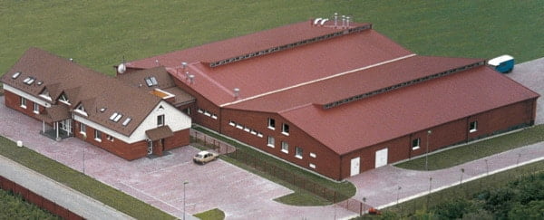 Luftaufnahme eines großen Gebäudekomplexes mit roten Dächern, bestehend aus mehreren verbundenen Hallen und einem kleineren angrenzenden Wohngebäude. Das Gelände ist umgeben von einer Zufahrtsstraße und gepflegtem Grün.