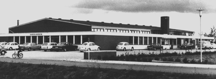 Schwarz-weiße Aufnahme eines flachen, rechteckigen Gebäudes mit mehreren großen Fenstern und einem Flachdach. Im Vordergrund befinden sich parkende Autos und ein Fahrrad, während eine Straße und Lampenlaternen das Bild rahmen.