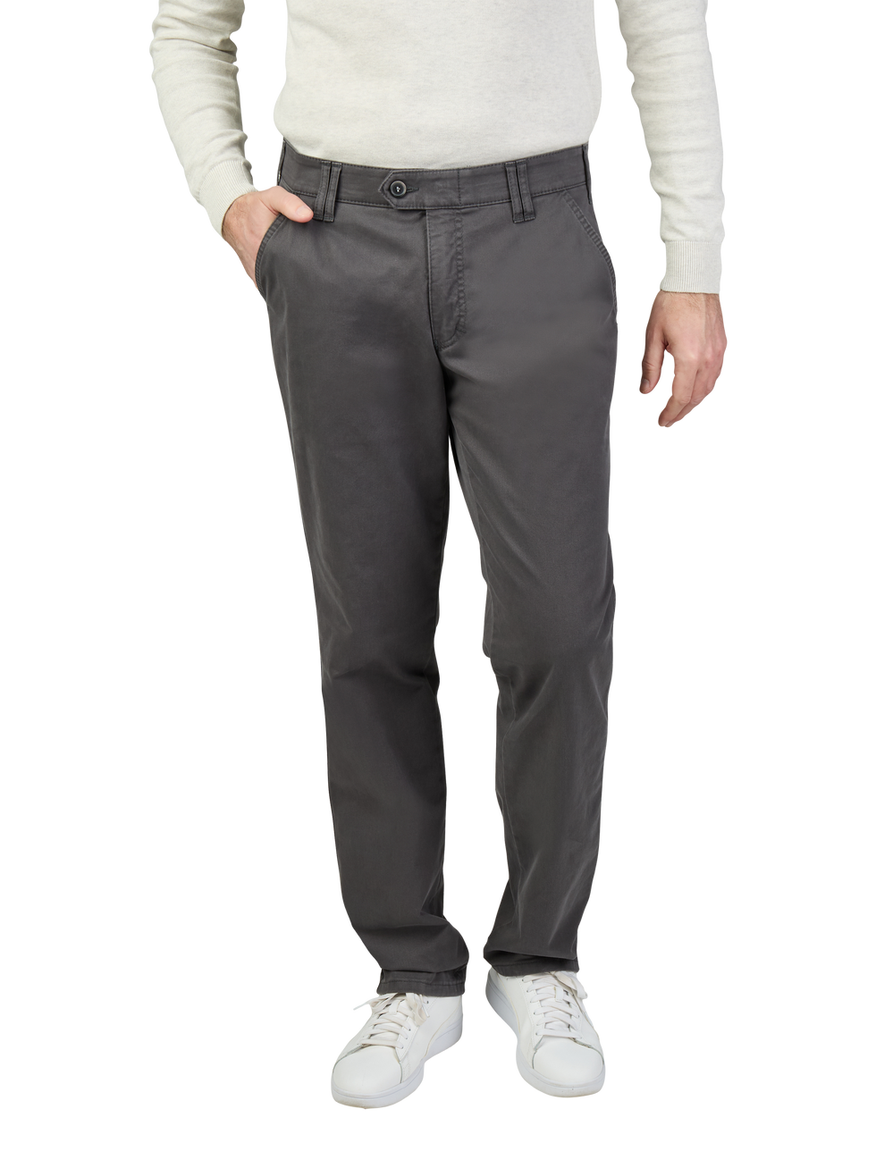 Mann trägt dunkelgraue, gerade geschnittene Chino-Hose mit Gürtelschlaufen und einer Seite mit Hand in der Tasche. Oberkörper trägt er ein hellbeiges, langärmeliges Oberteil, Schuhe sind weiße Sneaker.
