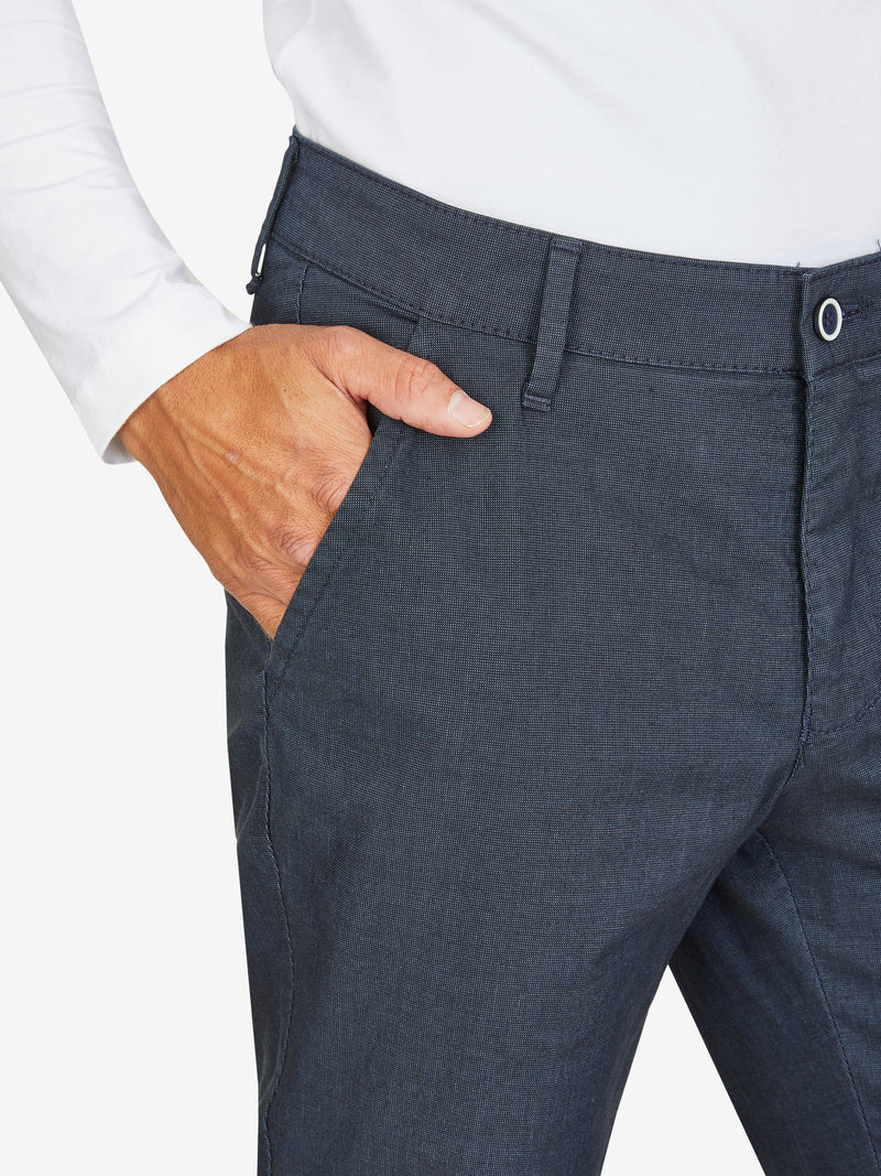 Nahaufnahme einer dunkelblauen, elastischen Flat Front-Hose mit Baumwoll-Mikrostruktur in Woll-Optik, mit Gürtelschlaufen und einer Hand im Bund. Modell zeigt Regular Fit, schmaler Beinverlauf, geeignet für Frühling/Sommer.