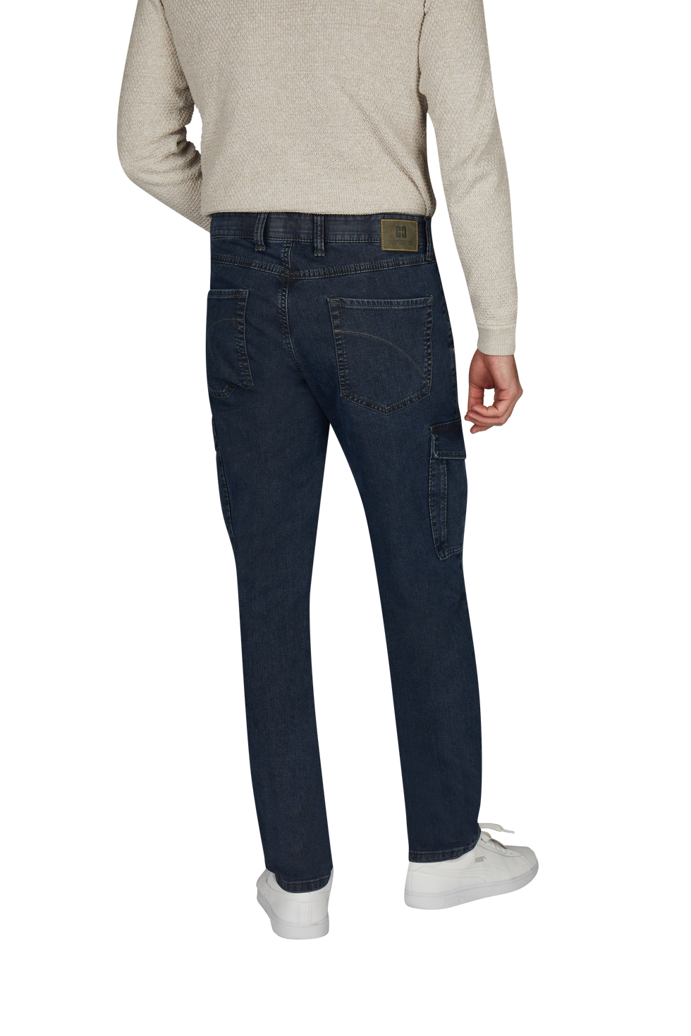 Ein Mensch steht mit dem Rücken zur Kamera und trägt eine beige, langärmlige Jacke, dunkle Jeans mit sichtbaren Nähten und Taschen, weiße Sneakers.