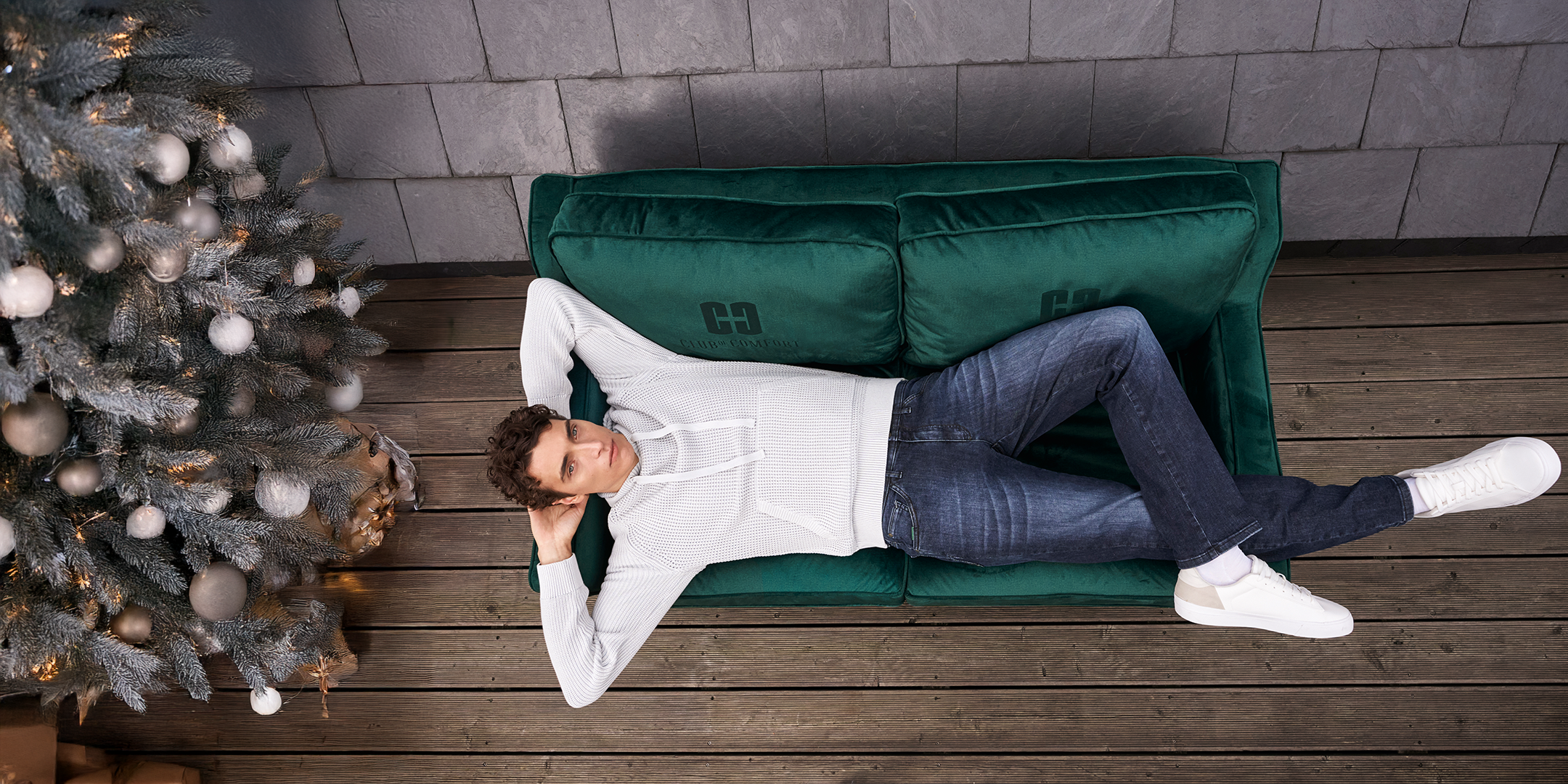 Ein Mann liegt entspannt auf einem grünen Sofa mit weißen Socken, trägt eine weiße Pullover und dunkle Jeans, während er mit geschlossenen Augen auf einer Seite liegt, neben einem Weihnachtsbaum mit silberner Dekoration, auf Holzfußboden vor einer grauen Ziegelwand.